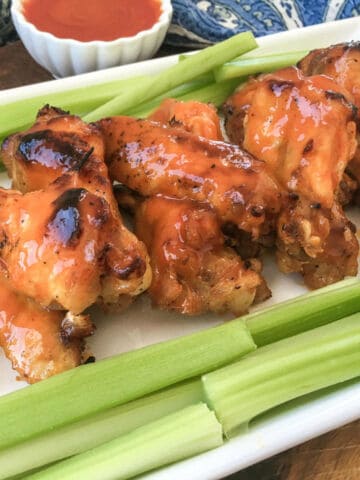 A plate of glazed Instant Pot buffalo chicken wings arranged in a row with fresh celery sticks, with a small bowl of red dipping sauce in the background.