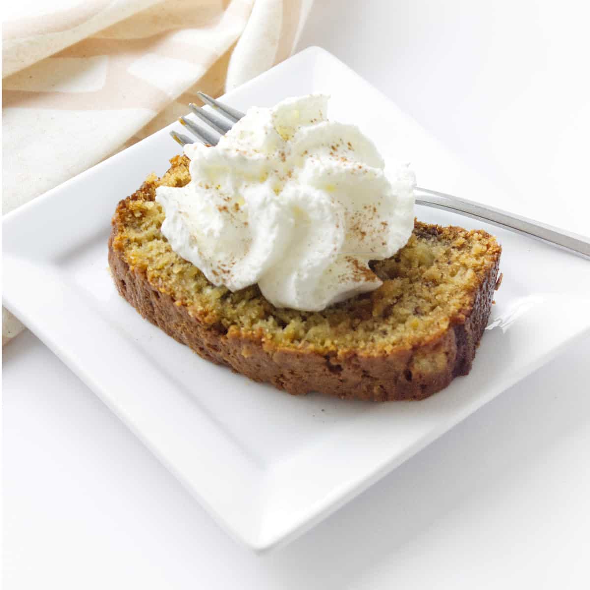 square white plate with fork and napkin holding slice of banana bread topped with whip cream.