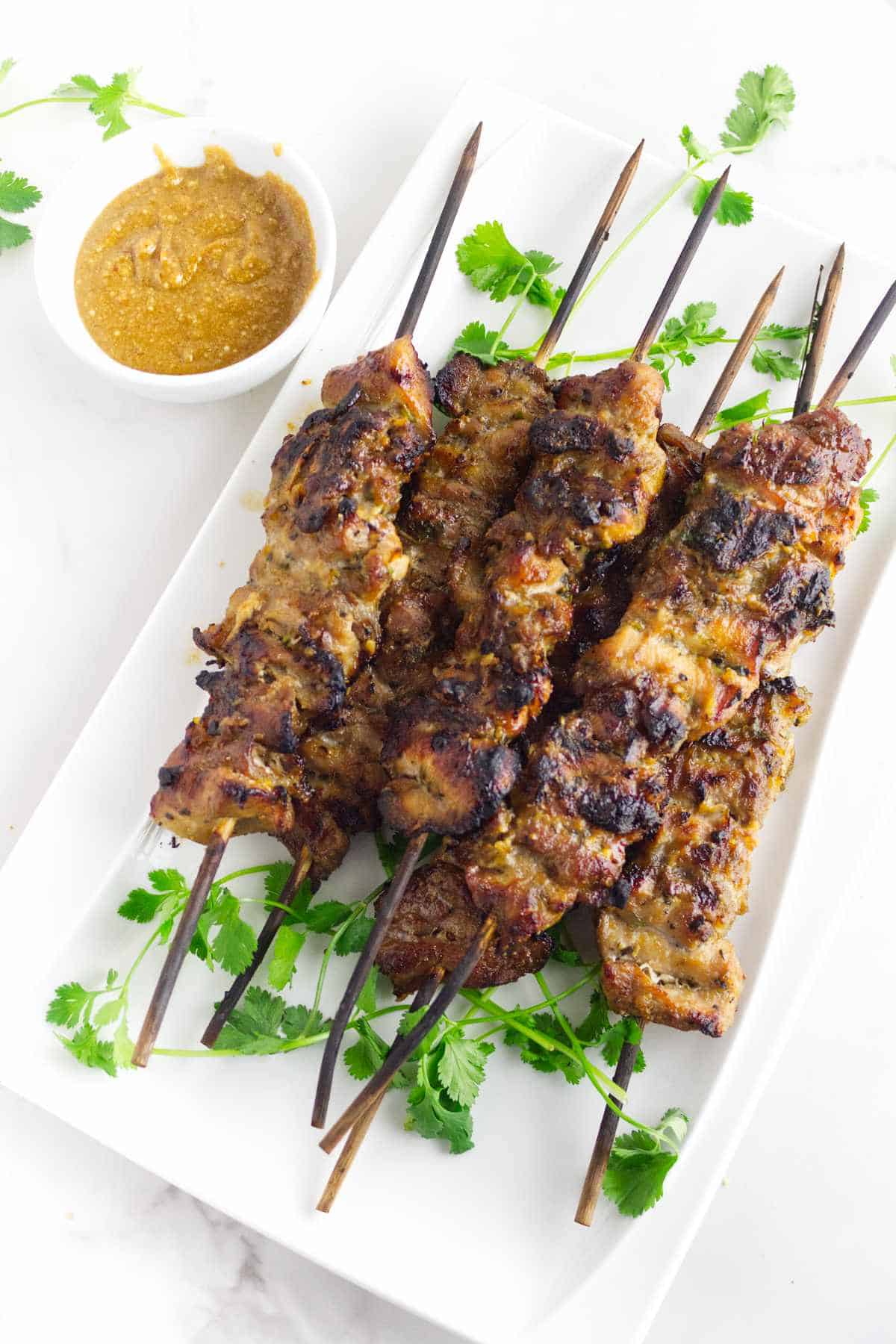 platter of grilled Thai pork skewers (moo ping) with cilantro garnish and spicy peanut dipping sauce.