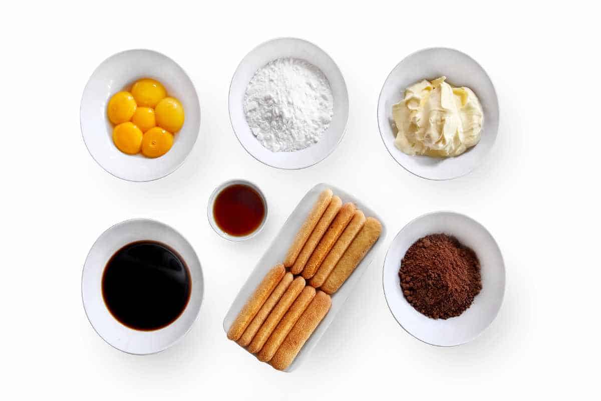 bowls holding egg yolks, powdered sugar, cream cheese, espresso, coffee liqueur, ladyfinger cookies, and cocoa powder.