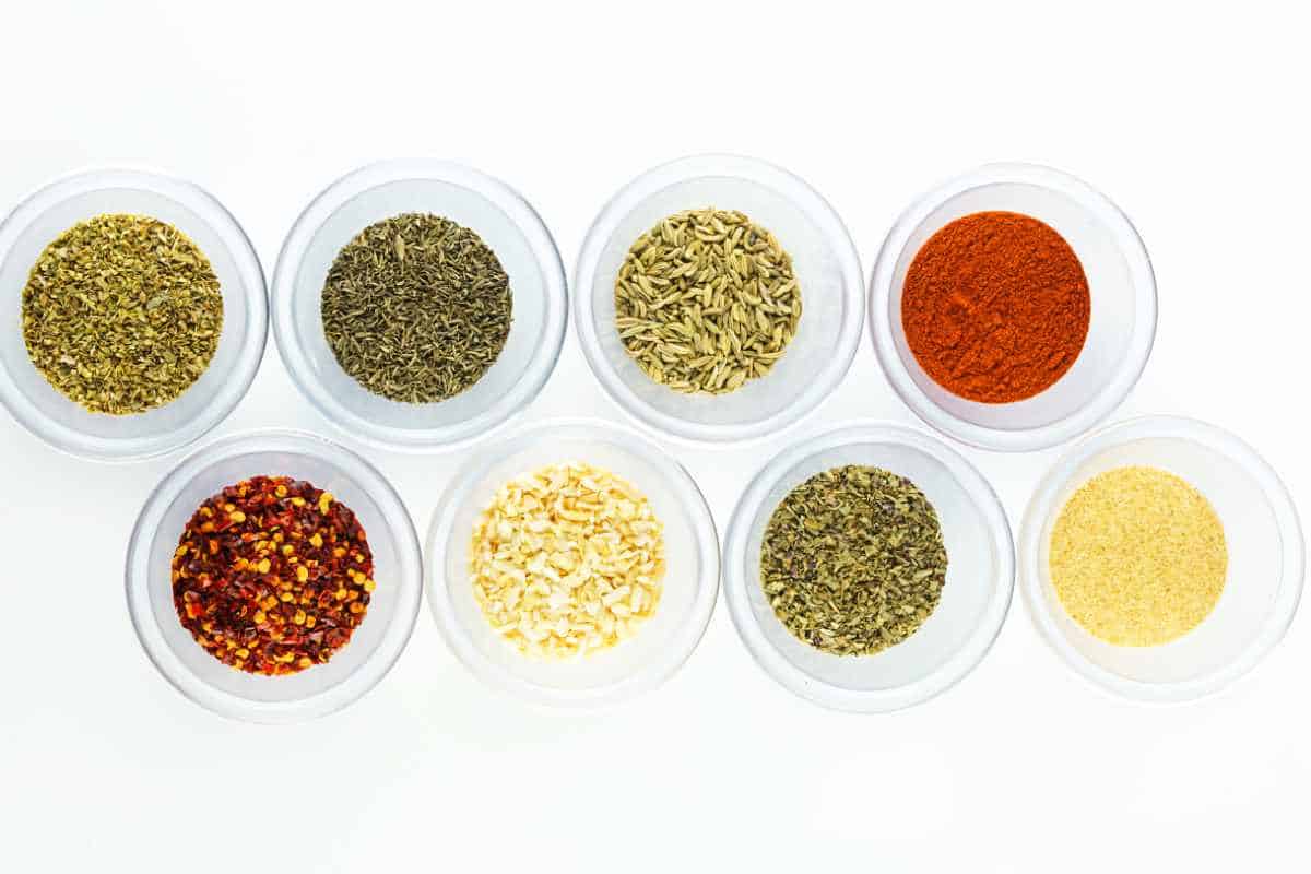 eight small bowls of colorful spices and herbs on a white background.