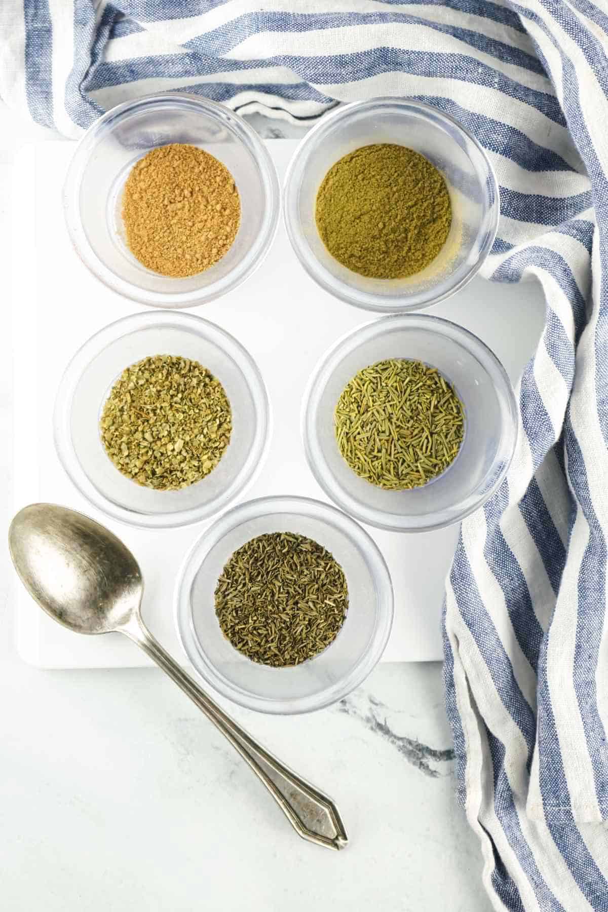 green herbs and spices in small bowls on a white background.