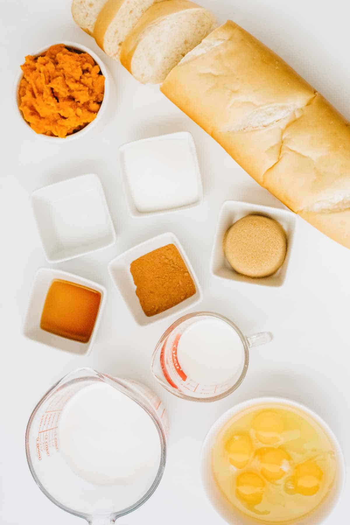 sliced french bread, pumpkin puree, sugar, spices, milk, and eggs on a white background.