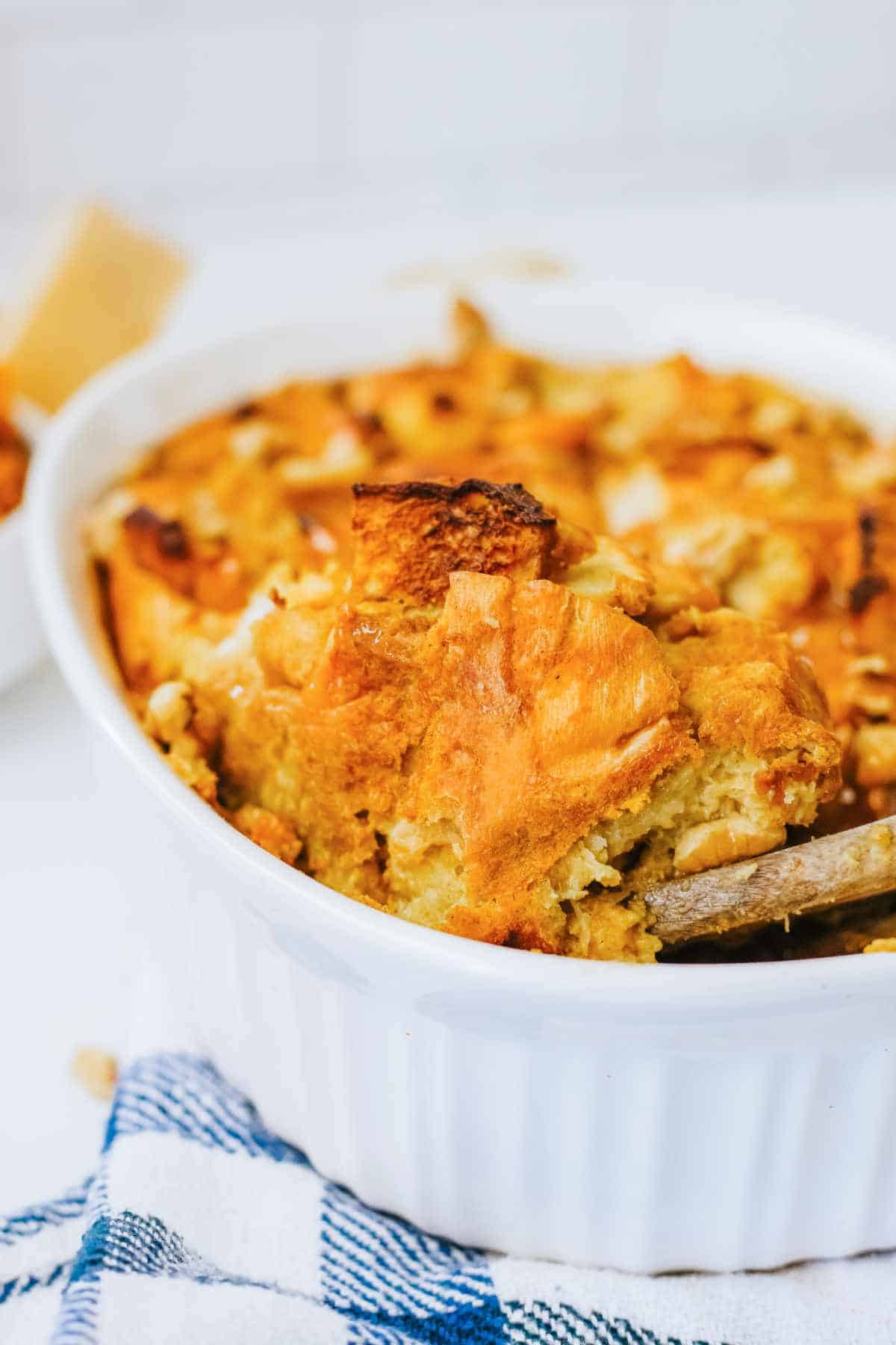 wooden scoop dishing up a serving from baked pumpkin bread pudding with nuts scattered on top in an oval white deep dish casserole.