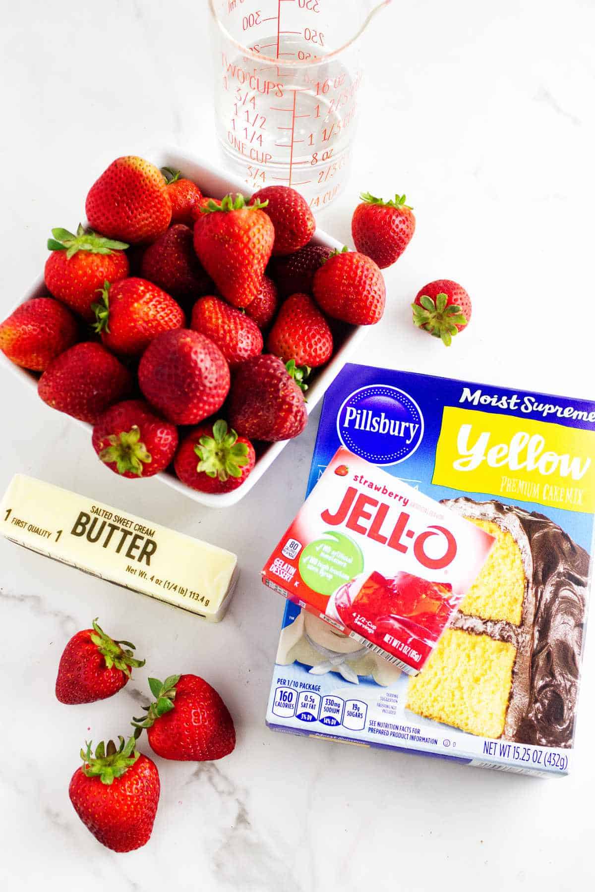 layout on a white background with a yellow cake mix, box of red jello, stick of butter, pitcher of water, and basket of strawberries.