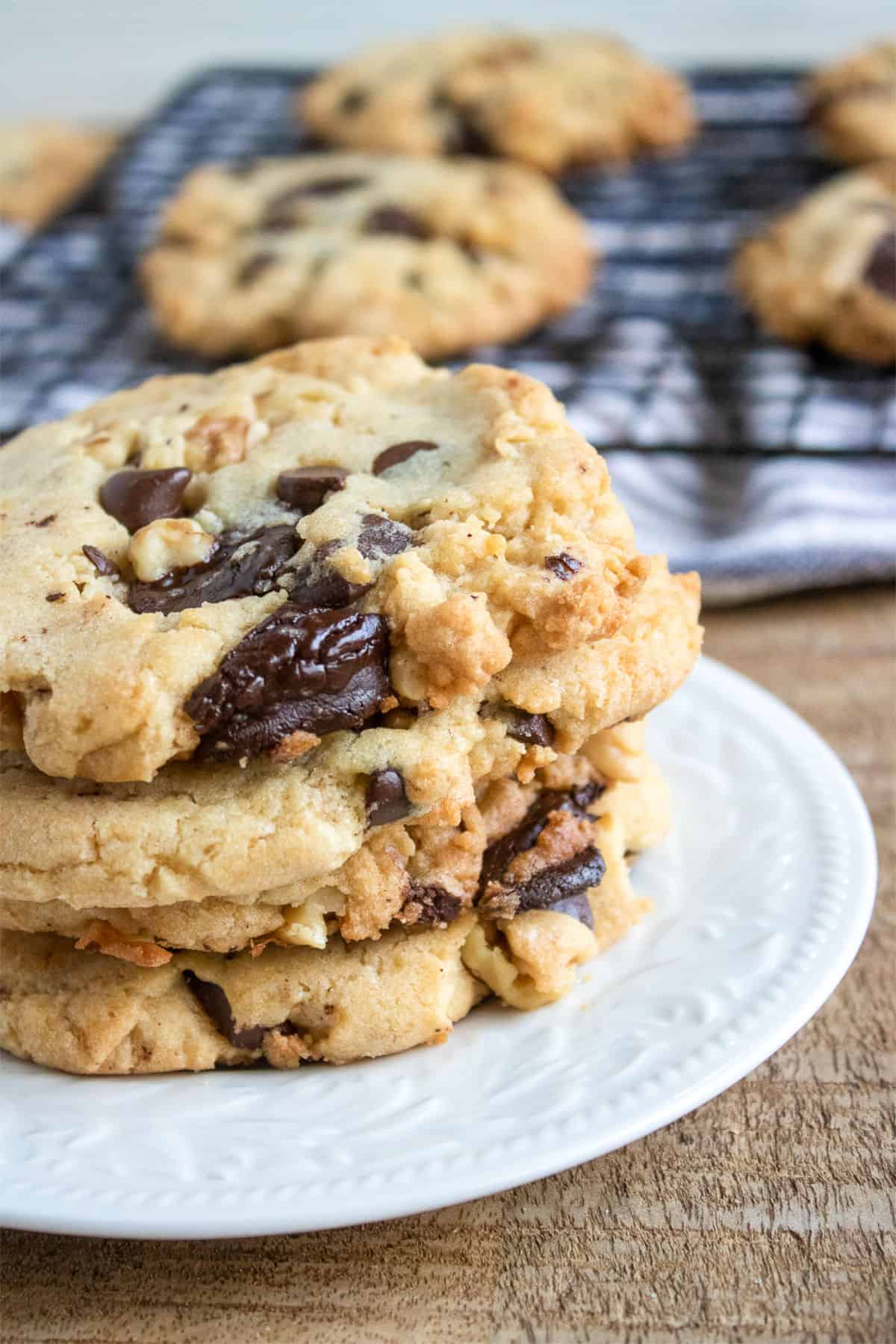 stack of four walnut chocolate chip cookies on a white plate with cooking cookies on a rack.