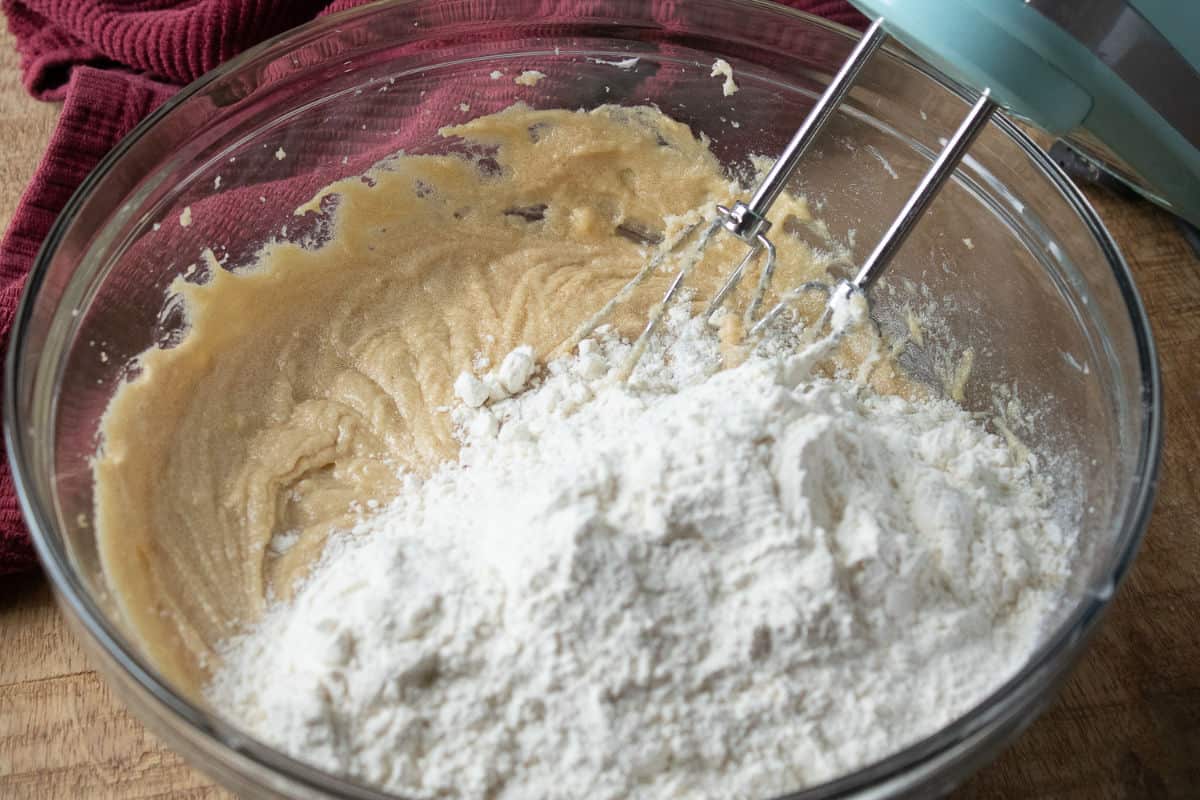 flour added to creamed sugar mixture in a clear glass bowl with mixer beaters in bowl.