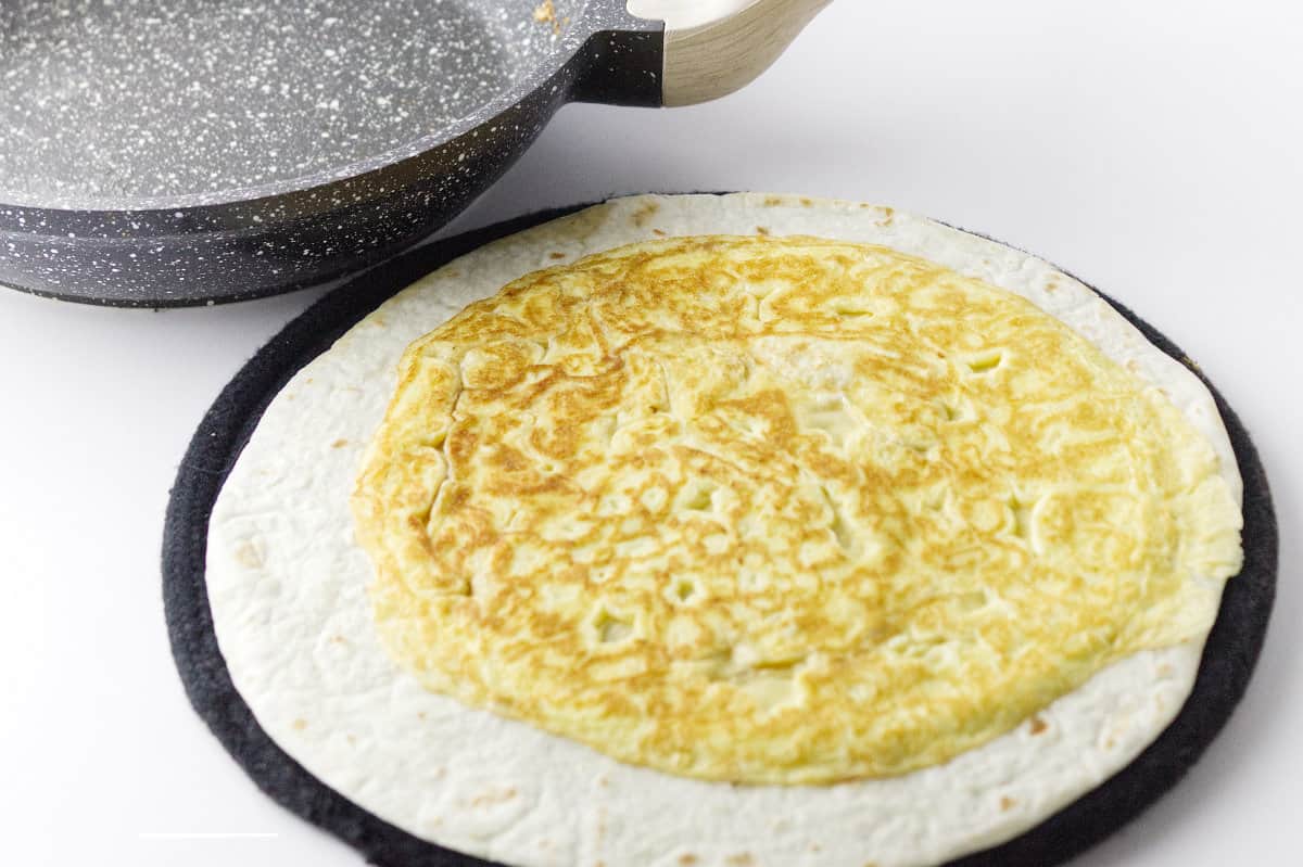 A flour tortilla on round board with a circle of egg on top with grey speckle skillet.