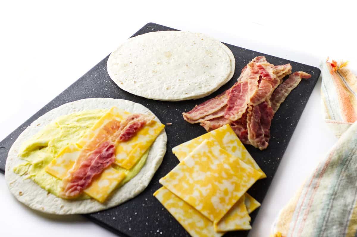 cutting board with Colby cheese, bacon strips, flour tortilla, and a loaded tortilla with egg, cheese, and bacon ready to roll up.