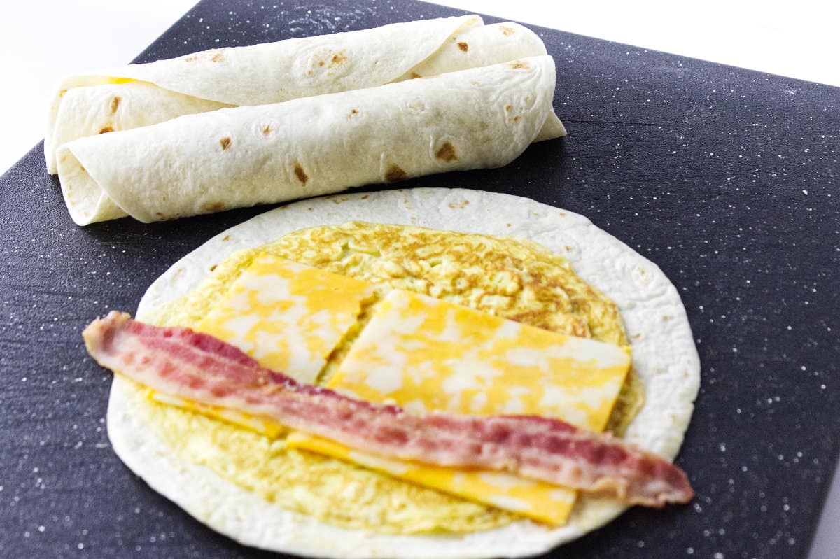 .cutting board with two rolled flautas and one flour tortilla laid out with egg, colby cheese and bacon to roll up into a flauta.