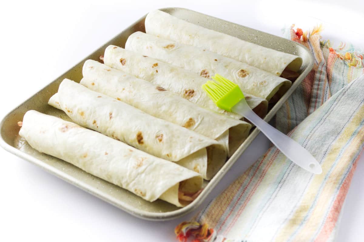 gold baking tray with six uncooked flautas, with yellow basting brush with butter.