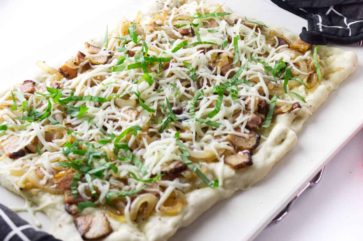 uncooked flatbread on a pizza stone with onion, bacon, mozarella and shredded basil.