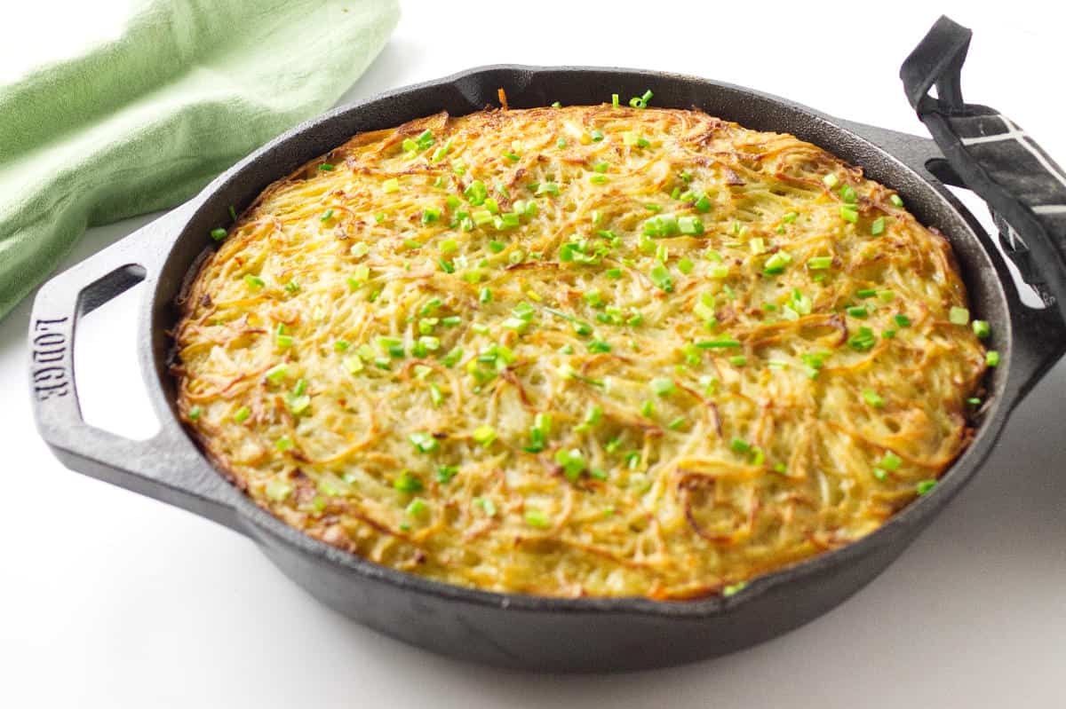 lodge cast iron skillet with grated potato kugel garnished with scallions on a white background and a green napkin and black pot holder