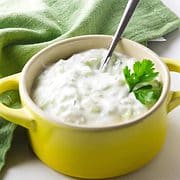 yellow ramkin with small spoon ladleing cucumer raita with a green napkin on a white background