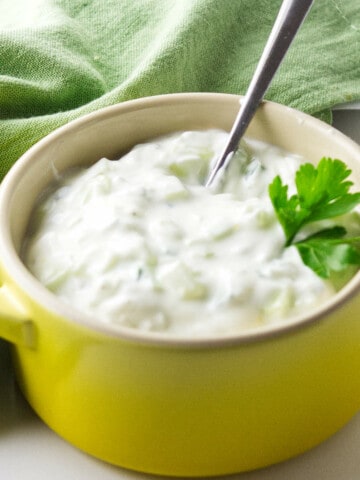 yellow ramkin with small spoon ladleing cucumer raita with a green napkin on a white background