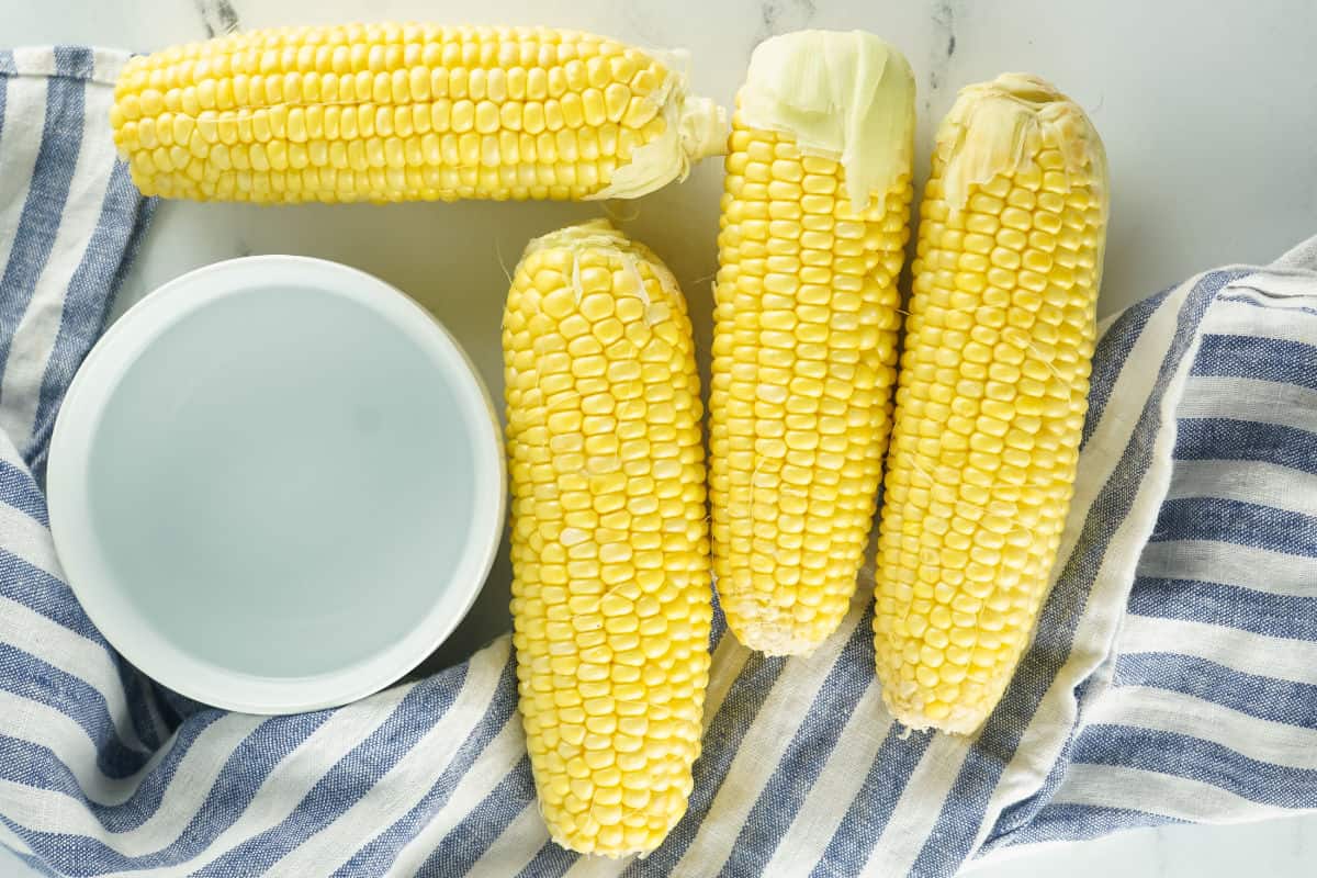 four ears of corn cleaned of silk on a white background with bowl of water and a striped napkin