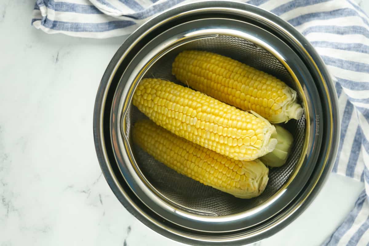 four ears of corn added to an Instant Pot with blue striped towel on a white marble background