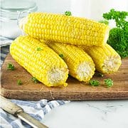ears of cooked corn on the cob on cutting board with knife, napkin, and parsley in background on white marble