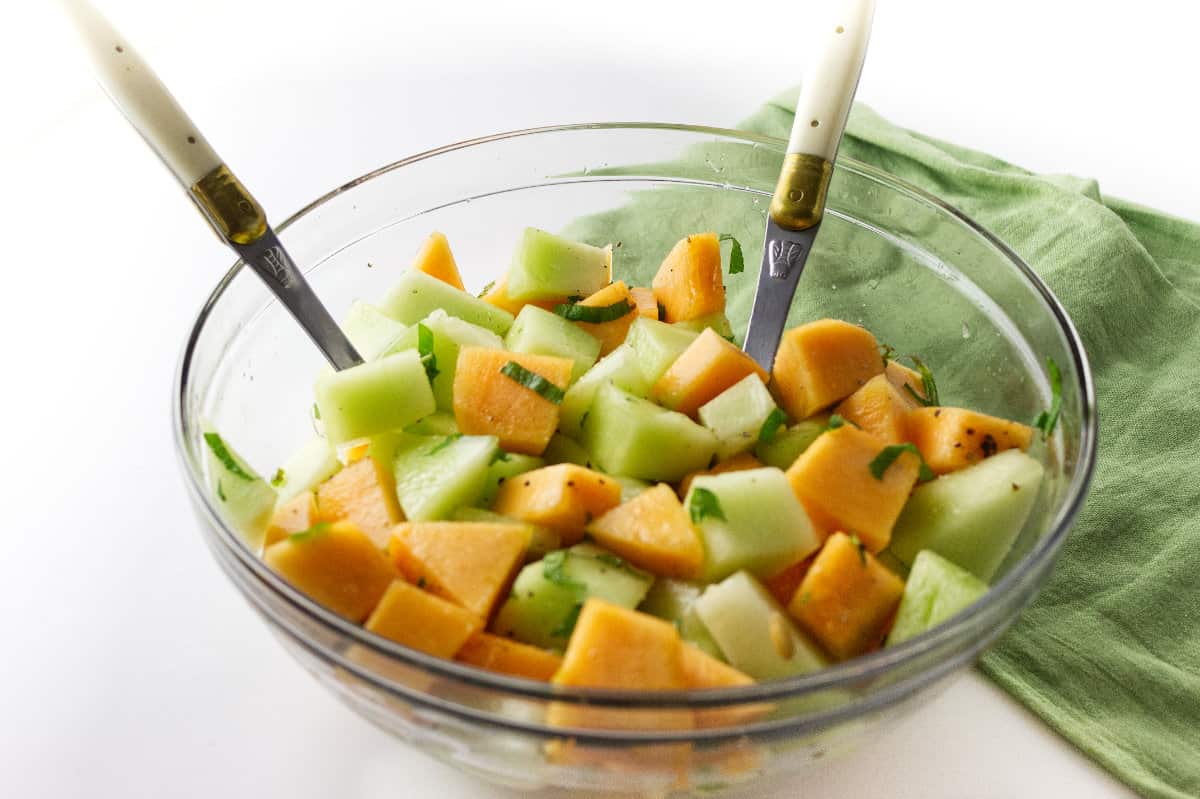 clear bowl with orange and green melon salad, salad spoon & fork with tossed dressing.