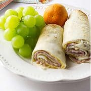 white plate with Monte Cristo Roll Ups, green grapes, and tangerine with orange stripe napkin on a white background