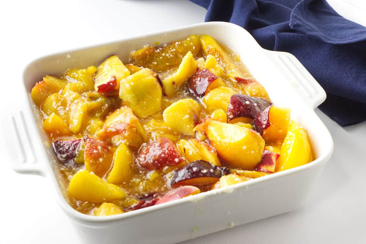 white baking dish full of sliced peaches, sugar and crumble ingredients.