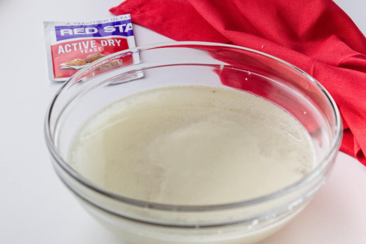 clear glass bowl with foamy yeast developing with yeast packet and red towel
