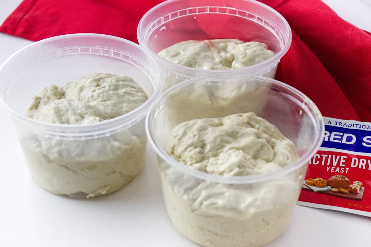 three plastic tubs with dough for the freezer with red star yeast packet and red towel on a white background