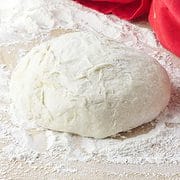 ball of dough rising on a floured wood board with red towel in background.