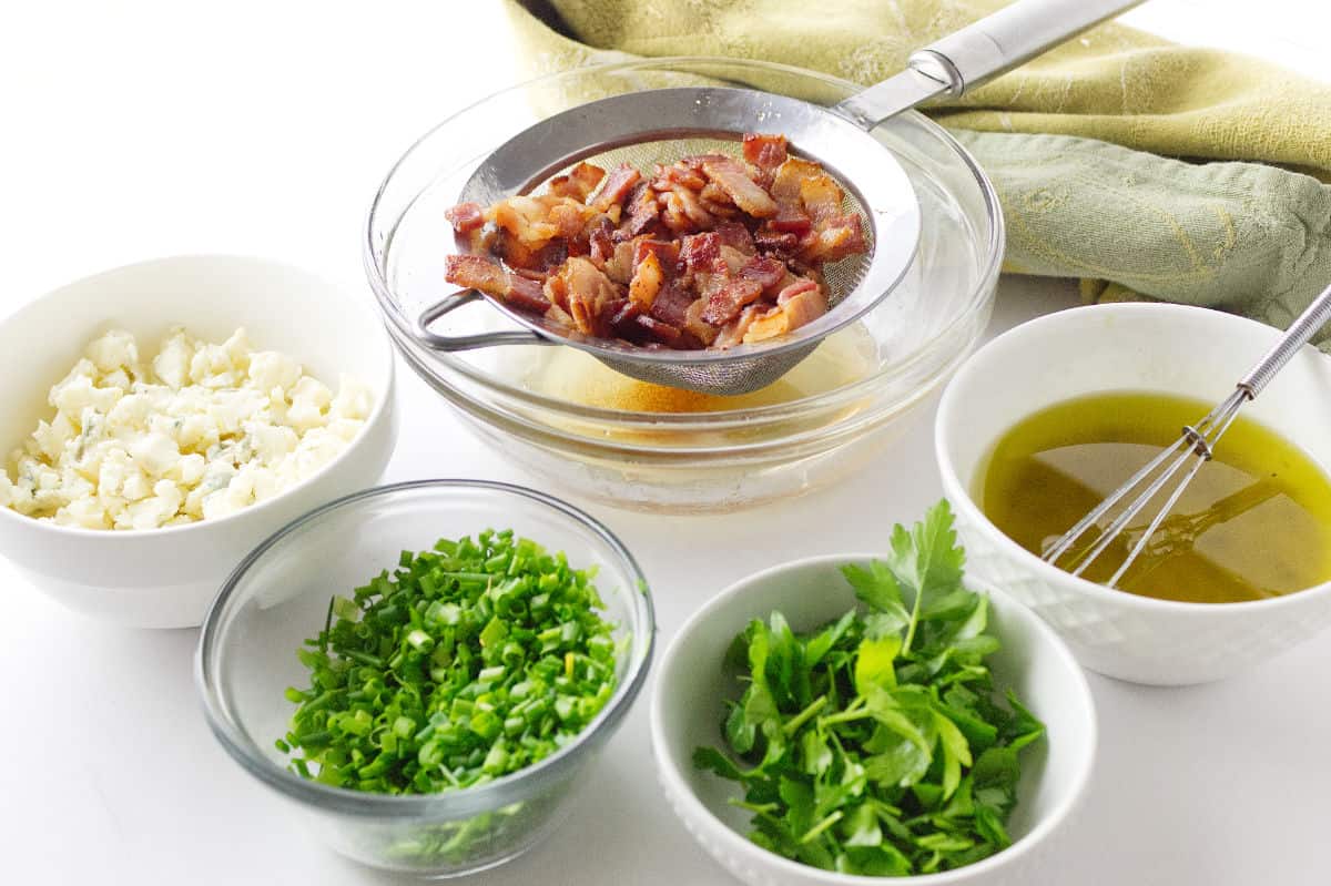 ingredients display for red hot and blue potato salad, bacon, blue cheese, scallions, parsley, olive oil in white bowls.