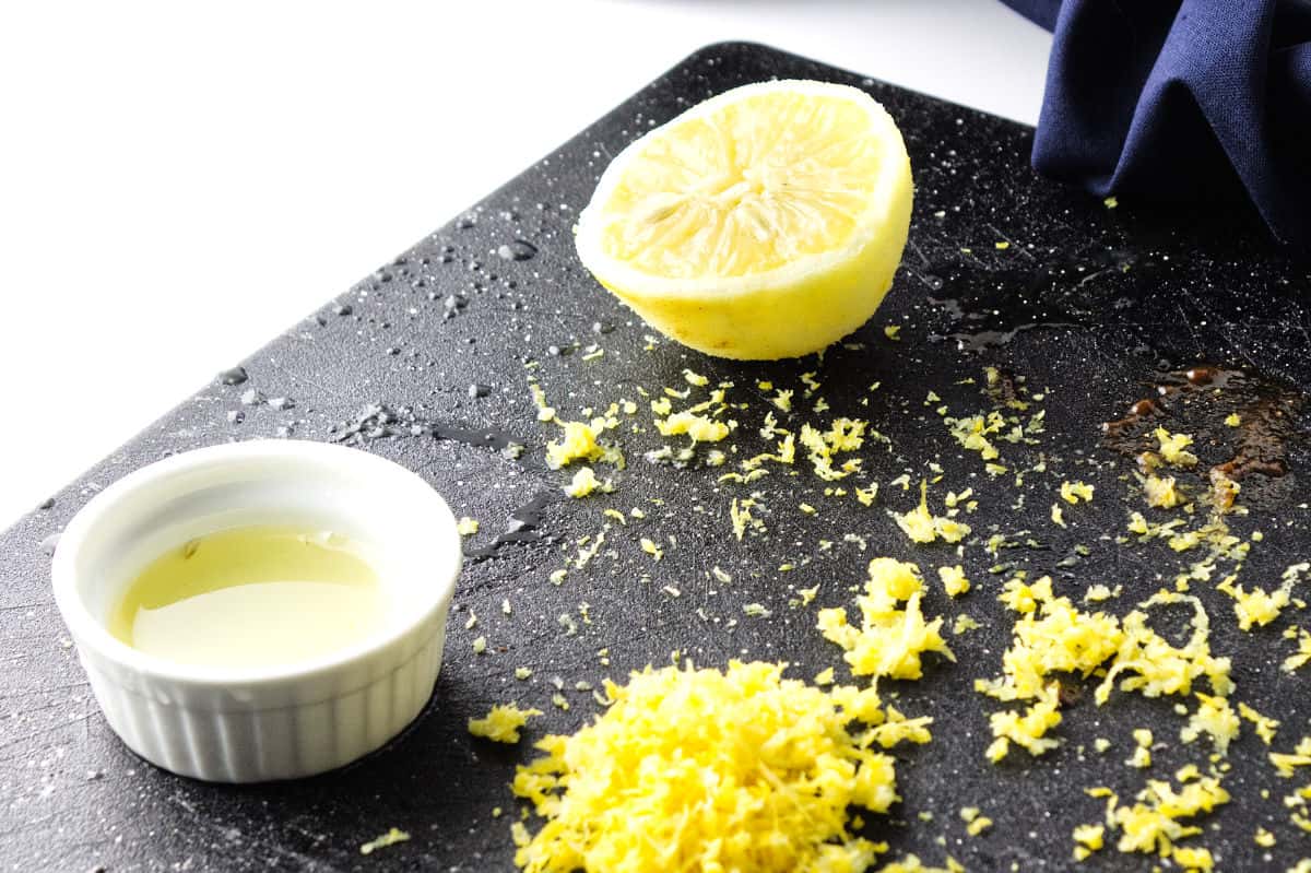 half a lemon with zest on a black cutting board and bowl of lemon juice nearby.