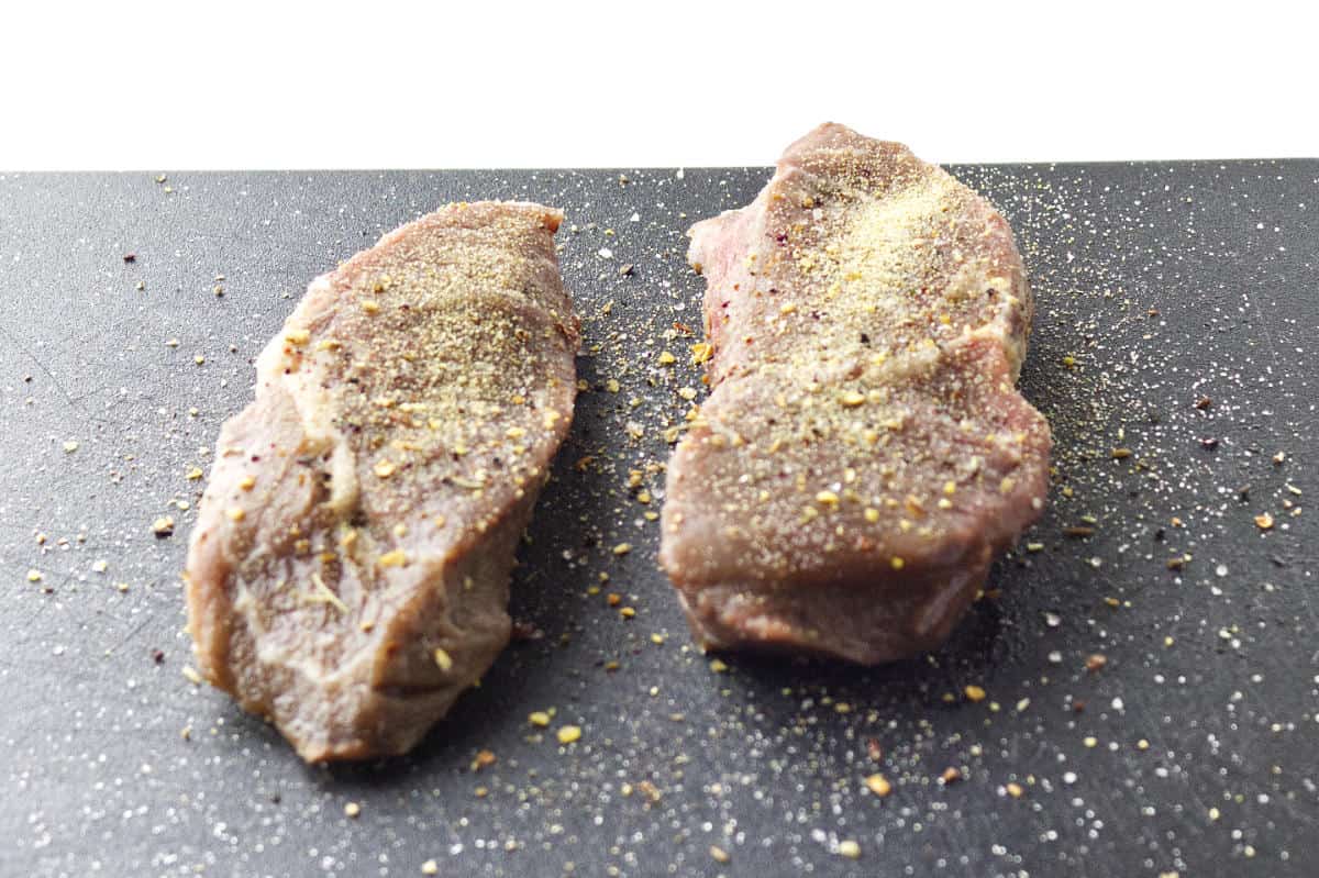 raw seasoned steaks on a black cutting board.
