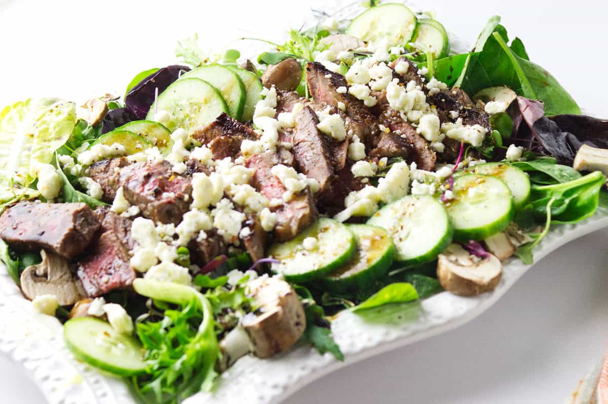 white serving platter with fresh lettuces and cucumber and slices of seared steak topped with feta cheese.