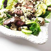 white serving platter with fresh lettuces and cucumber and slices of seared steak topped with feta cheese