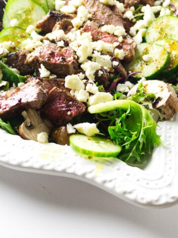 white serving platter with fresh lettuces and cucumber and slices of seared steak topped with feta cheese