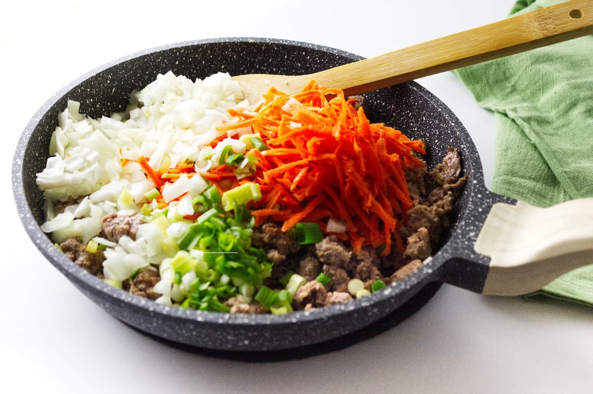 grey speckled skillet with ground pork, carrots, scallion, onion and wooden spoon.