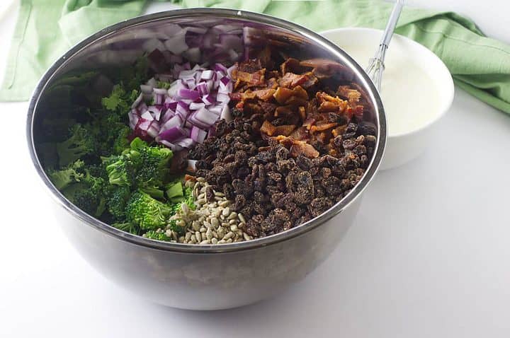 Metal bowl with kale, broccoli, onion, seeds, raisins, bacon; creamy dressing and green cloth in background.