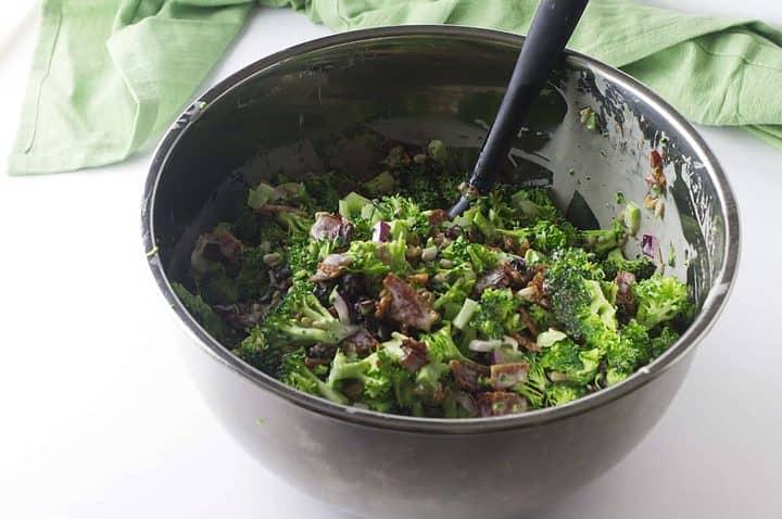 Metal bowl of Walmart broccoli salad with bacon, red onions, creamy dressing, black spoon, and green cloth in background.