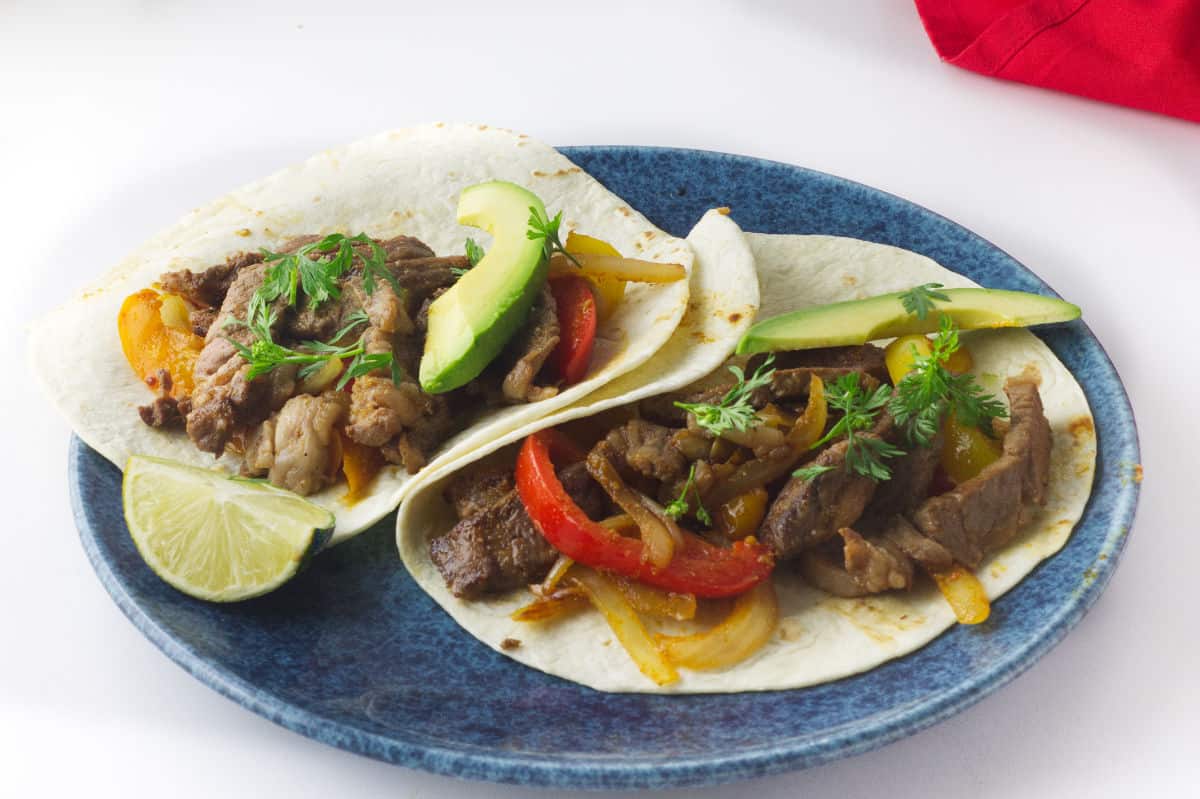 blue plate with flour tortillas loaded with sliced steak, onions, and peppers to roll up as fajitas.