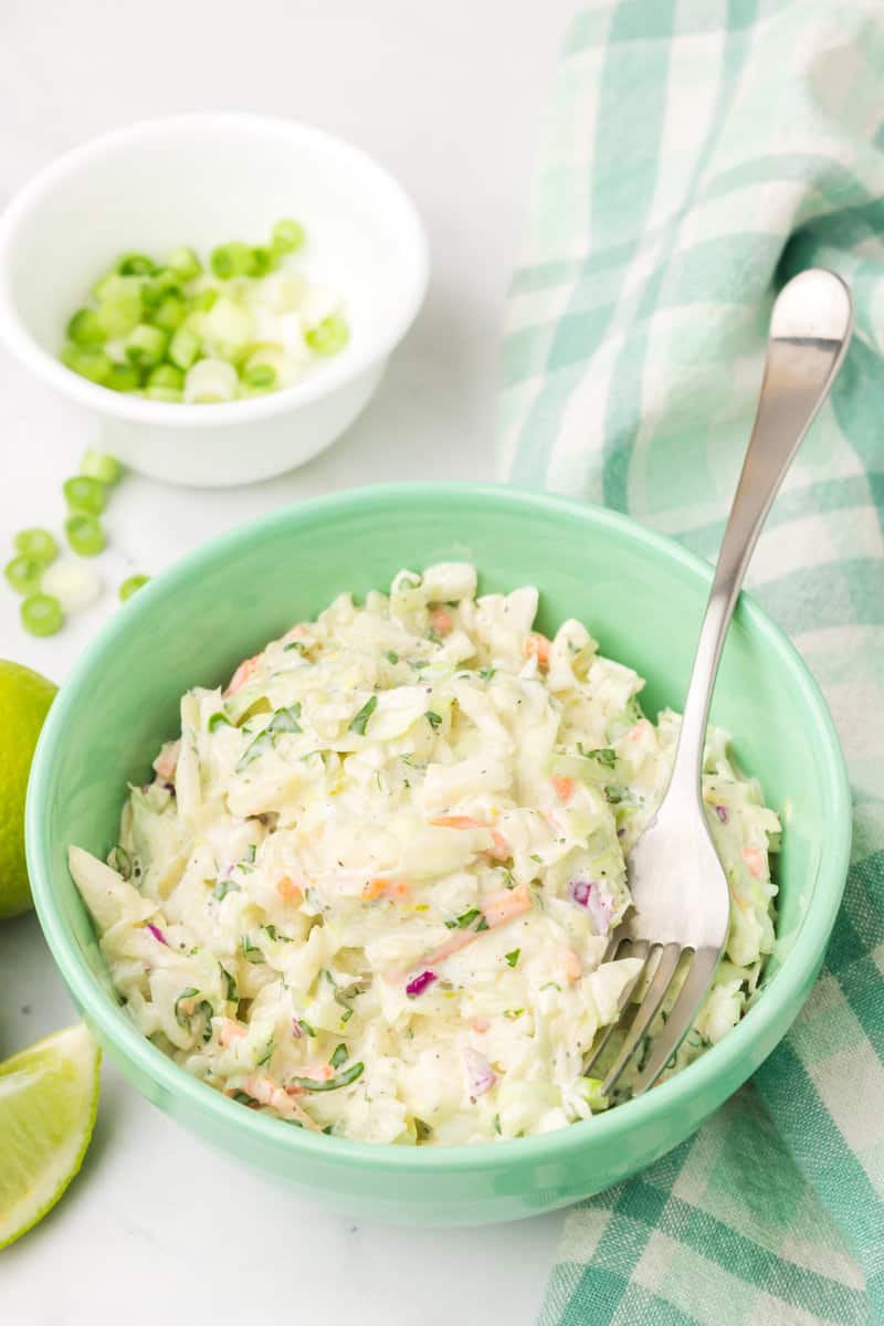 bowl of coleslaw with limes and green onions nearby.