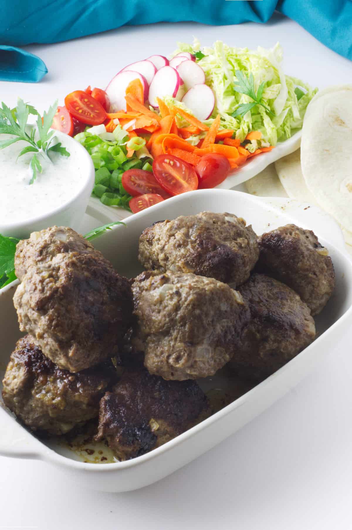 cooked lamb kofta balls in a serving dish with wraps, vegetables, and Tzatziki sauce.
