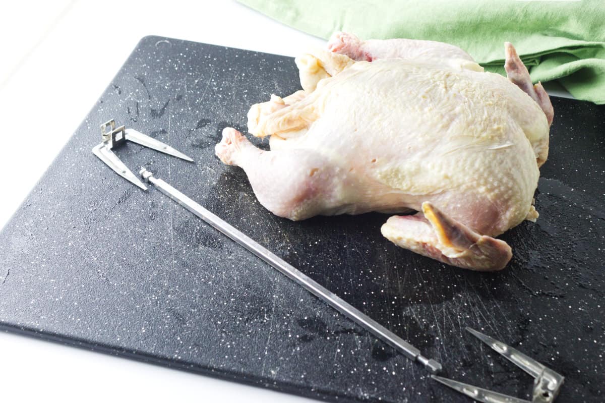 whole raw chicken fryer with rotisserie spit resting on a black cutting board