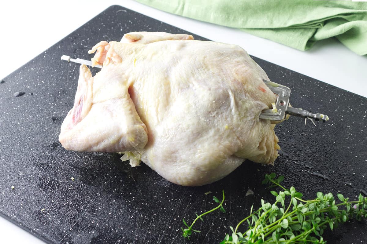 whole chicken fryer trussed up and on a rotisserie spit with fresh herbs on the cutting board