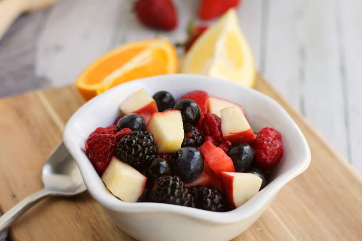 brown wood cutting board with white bowl of cut fruit salad with sliced oranges in background.