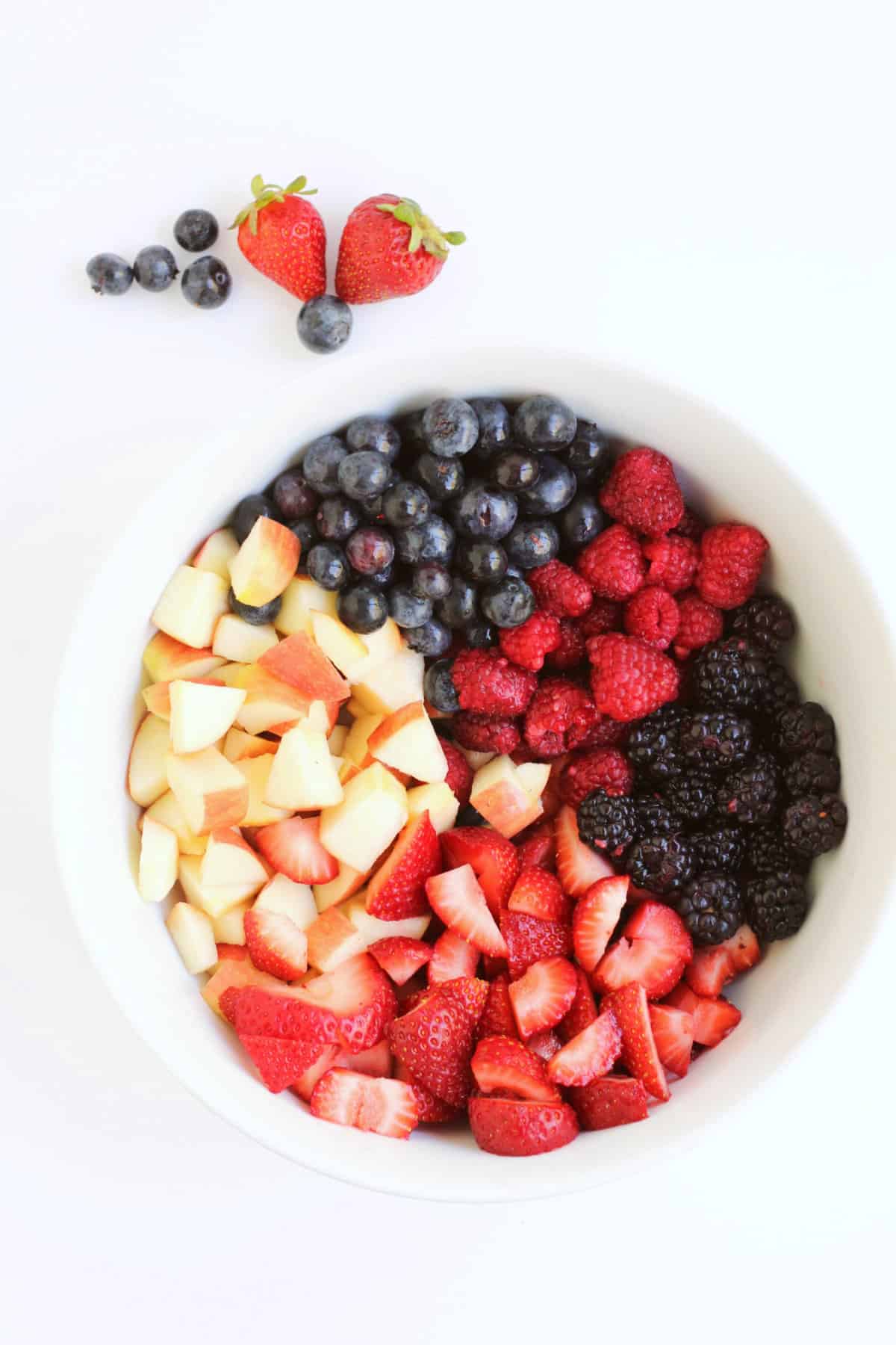 white bowl of diced apples and strawberries, and whole blueberries, raspberries, and blackberries.