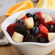 white bowl of summer fruit salad of berries and diced apples with oranges and strawberries in the background