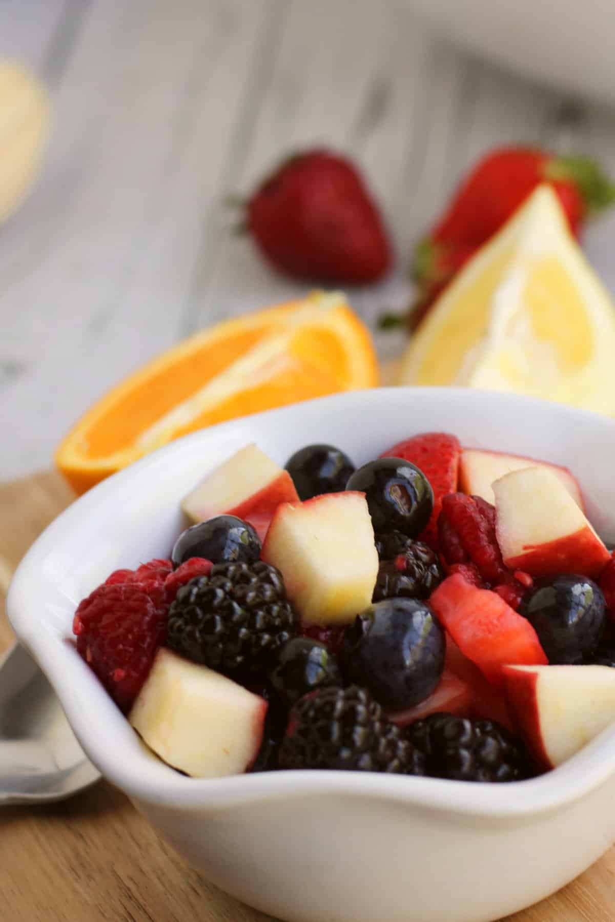 white bowl of summer fruit salad of berries and diced apples with oranges and strawberries in the background