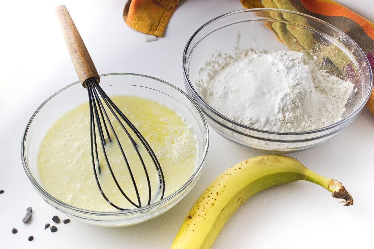 a bowl of wet ingredients and a bowl of flour and sugar with banana on counter.