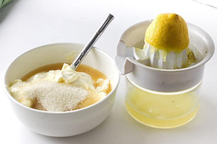 White bowl mixing mayo, sugar, and liquid sits next to a lemon juicer with a squeezed lemon half and juice below.