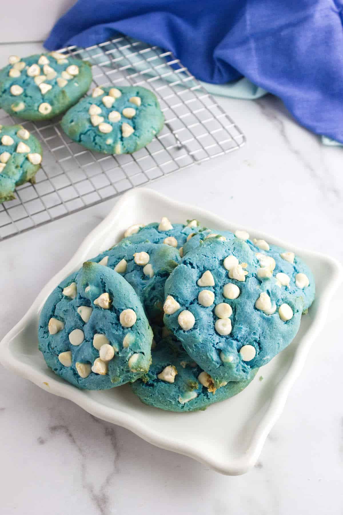 Four blue velvet cookies with white chips on a dish; more cookies, a cooling rack, and a blue cloth on a marble surface nearby.