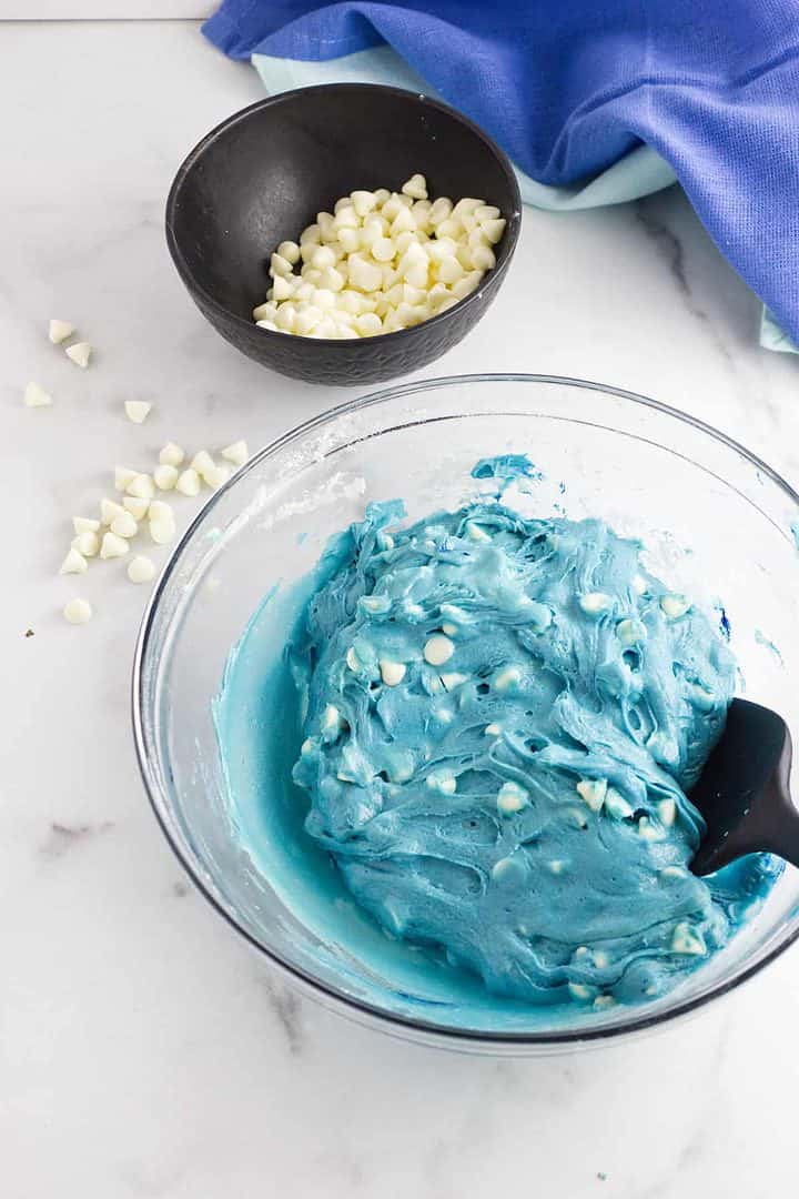 Bright blue velvet cookie dough with white chocolate chips in a glass bowl, black spatula, chips bowl, and blue cloth nearby.